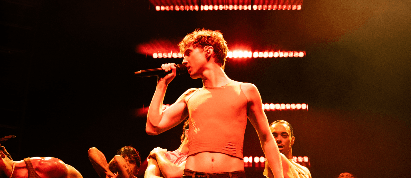 A person in a sleeveless shirt sings into a microphone on stage, surrounded by dancers with illuminated lights in the background.