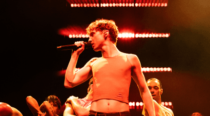 A person in a sleeveless shirt sings into a microphone on stage, surrounded by dancers with illuminated lights in the background.